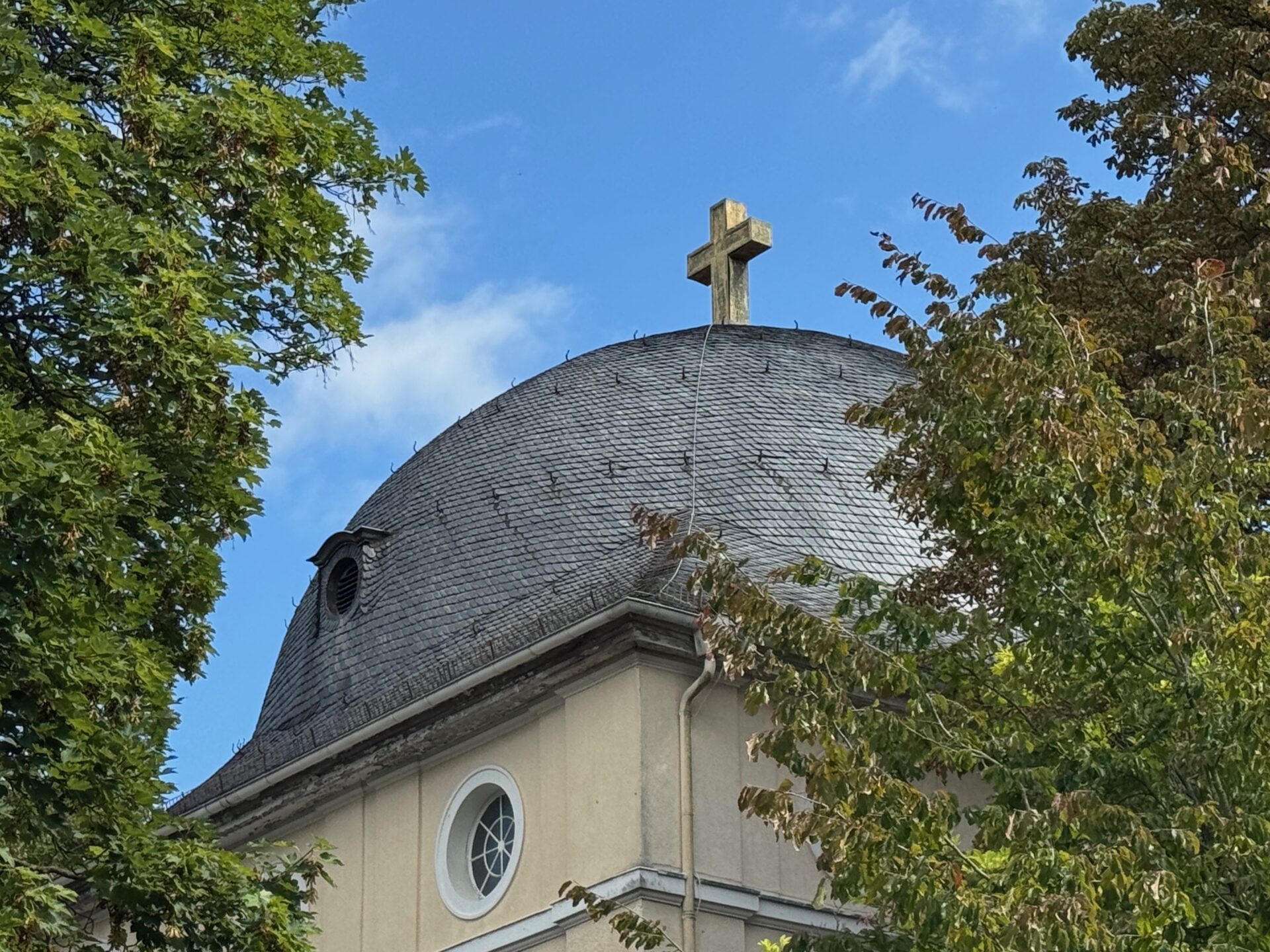 Das kuppelförmige Kirchendach der Trauerhalle des Friedhof Höchst in Frankfurt am Main mit einem Kreuz darauf, das teilweise von grünen Bäumen verdeckt wird, erhebt sich vor einem blauen Himmel.