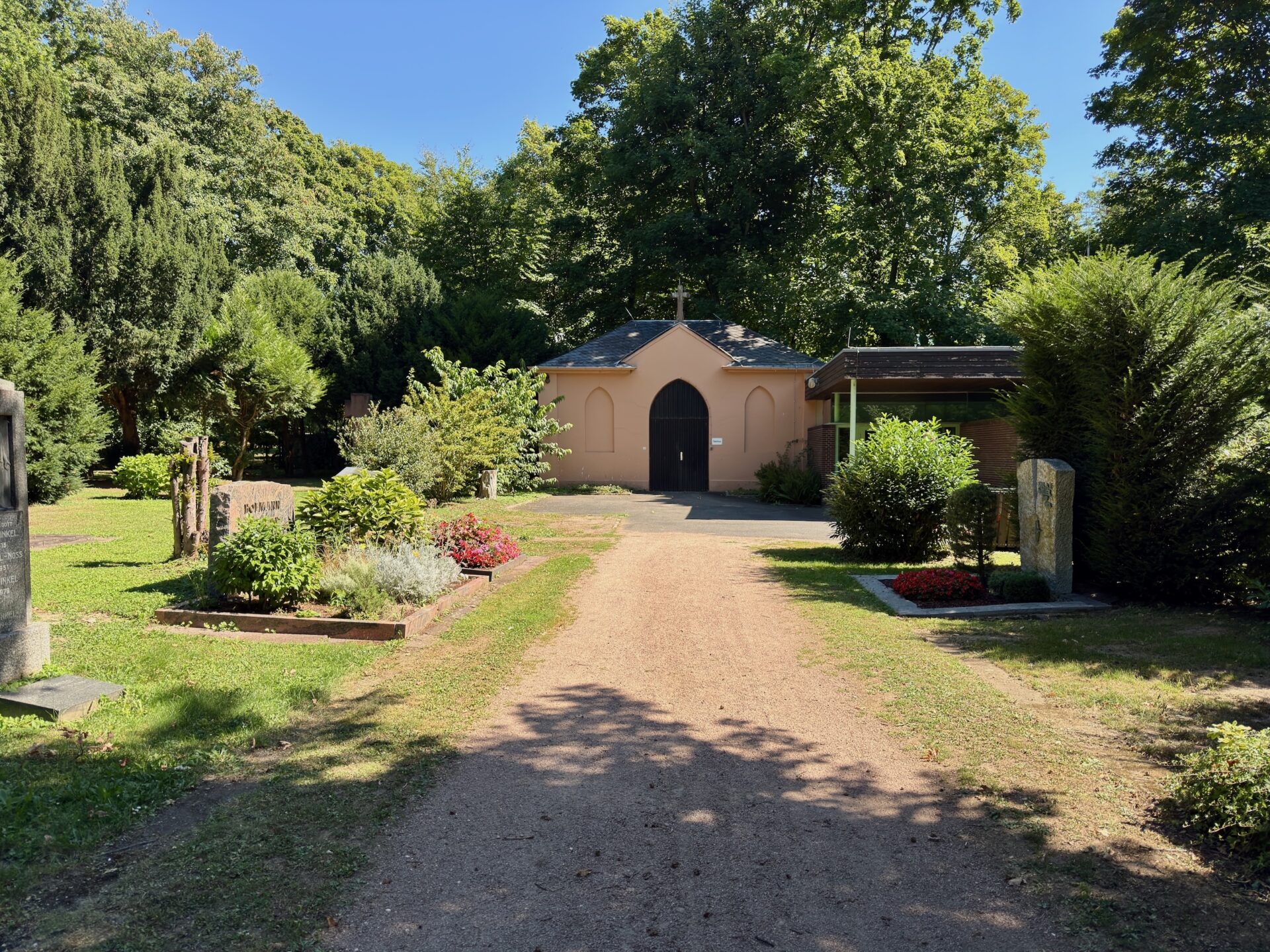 Ein sonnenbeschienener Gartenweg führt zur Totenhaus-Kapelle mit gewölbten Türen auf dem Friedhof Sossenheim in Frankfurt am Main, umgeben von Bäumen, Sträuchern, Blumenbeeten und Grabsteinen unter einem klaren blauen Himmel.