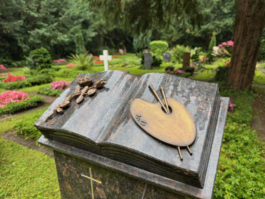 Grabsteinkunst eines aufgeschlagenen Buches mit einer Farbpalette und Pinseln, eingebettet in den üppigen, farbenfrohen Hauptfriedhof Frankfurt am Main, umgeben von Grabsteinen, Blumen, Bäumen und einem weißen Kreuz im Hintergrund.