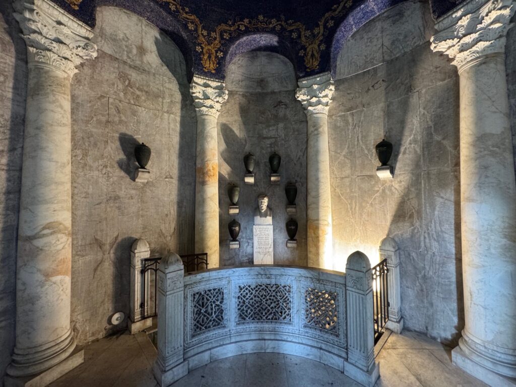Das Innere des Gans Mausoleums auf dem Hauptfriedhof Frankfurt am Main mit einem halbrunden Geländer, einer zentralen Büste, sechs schwarzen Urnen an der Wand dahinter, hohen Säulen und einer blau-goldenen, verzierten Decke. Warmes Licht hebt das weiße Mauerwerk hervor.